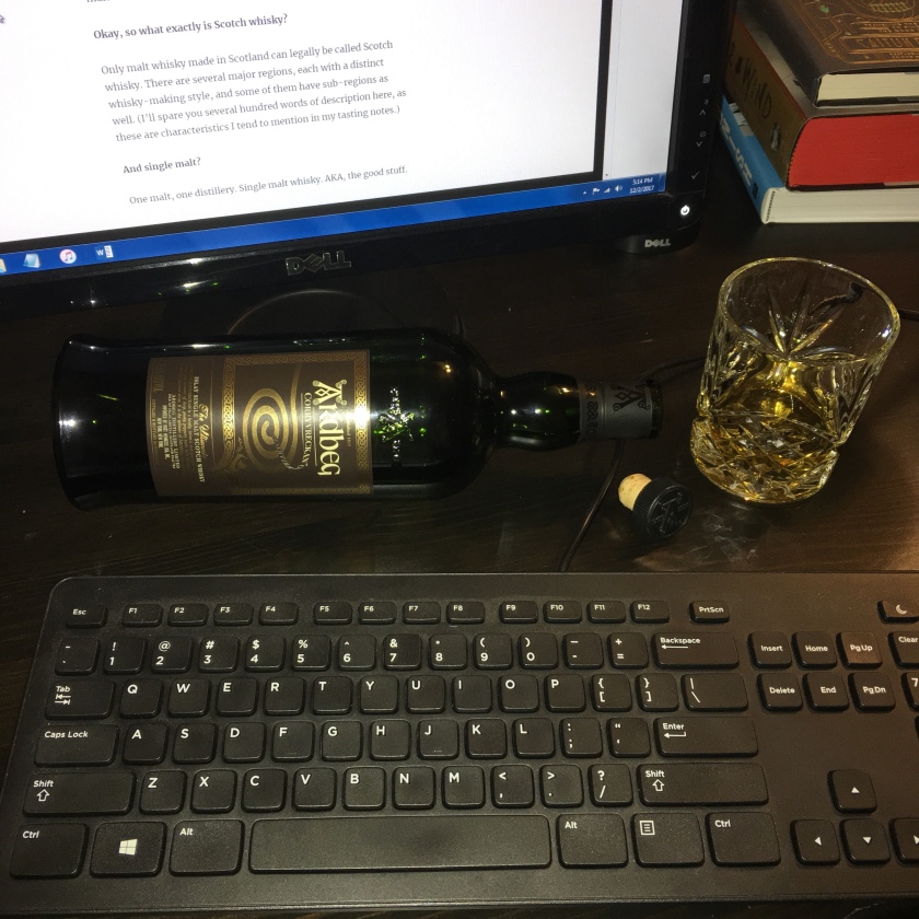 A bottle of Ardbeg Corryvreckan lies on its side, apparently empty, its cork a filled whisky glass nearby. These items are arranged near a keyboard and a computer monitor. On the monitor is writing about whisky. 