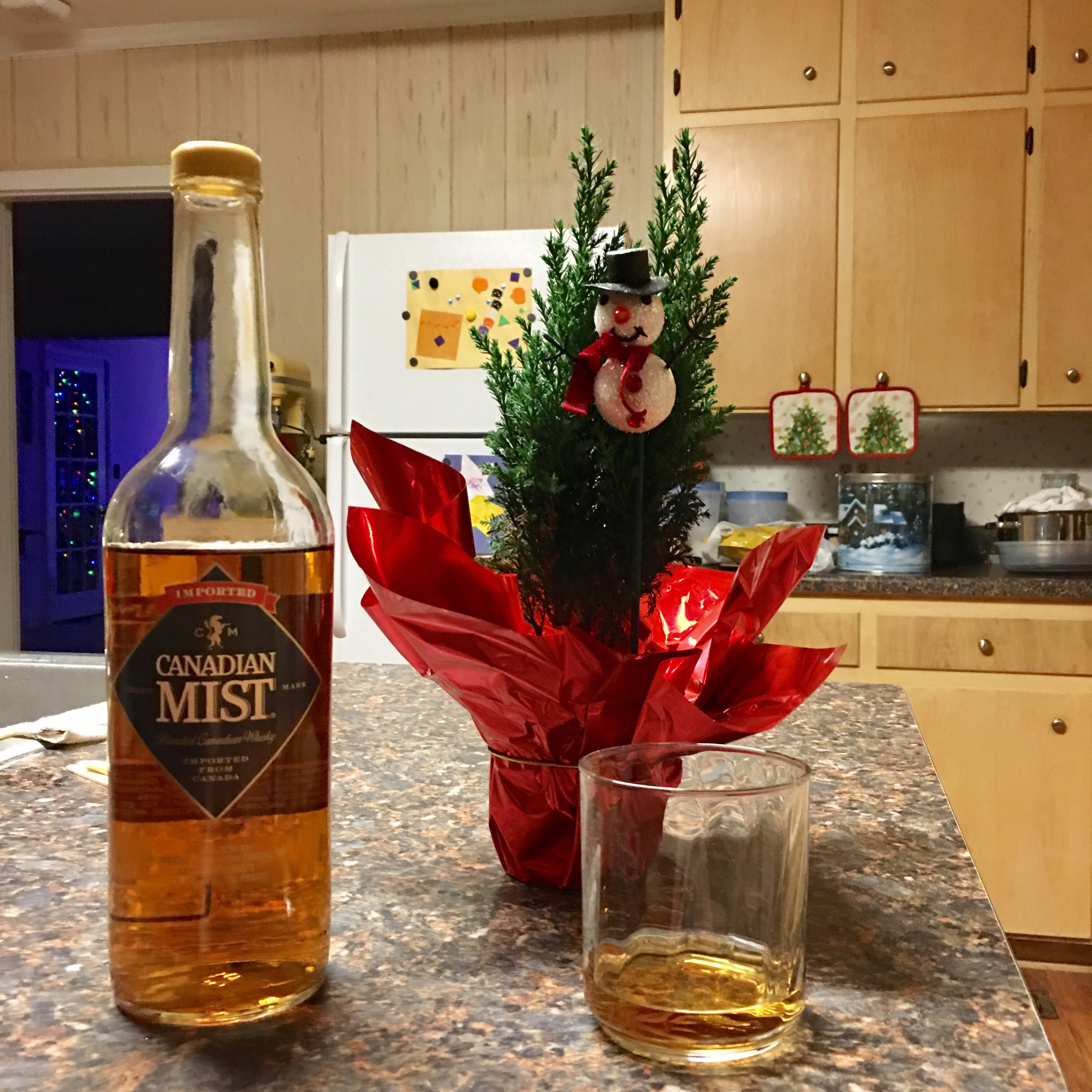 A bottle of Canadian Mist whisky sits next to a filled glass, next to a tiny tabletop Christmas tree. All of these items sit upon a kitchen counter. 